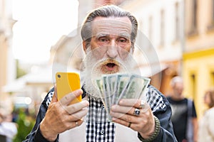 Happy elderly man counting money dollar cash, use smartphone calculator app in urban city street