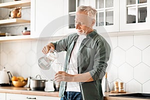 Happy elderly man filling his glass with water from jug in kitchen