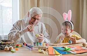 Happy elderly man granfather preparing for Easter with grandson. Portrait of smiling boy with bunny ears painted  colored eggs for