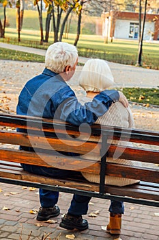 Happy elderly man and woman sitting on a bench in autumn day. Relaxed senior couple sitting on a park bench. Grandfather gently