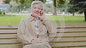Happy elderly woman with beautiful face sitting on park bench takes phone call, gray-haired lady of retirement age