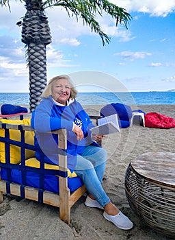 Happy elderly woman in blue clothes, sitting in a chair on the beach, looking at the camera, hold a book with a background of sea