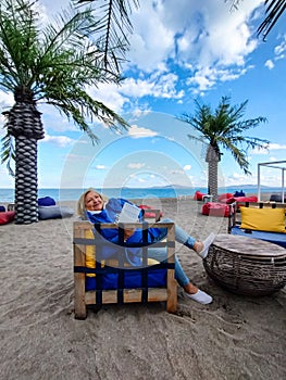 Happy elderly woman in blue clothes, sitting in a chair on the beach, reading a book with background of sea and palm