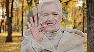 Happy elderly woman joyfully smiling standing in autumn park mature grandmother looking at camera showing gesture okay