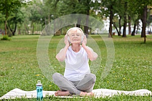 happy elderly woman listening music with headphones while sitting on yoga mat with bottle of water