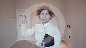 Happy elderly woman realtor shows the keys to the real estate to the camera on the background of an empty apartment for