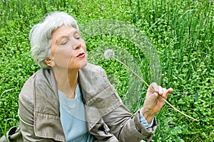Happy elderly woman sitting on a meadow blowing on a dandelion