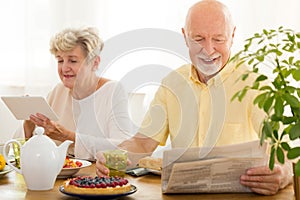 Happy elderly woman using a tablet and her husband reading a new