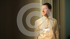 Happy elegant groom getting ready for the wedding day in the hotel room. Thoughtful man with eyes closed lit by lamp