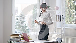 Happy elegant slim businesswoman in headphones dancing in office indoors. Portrait of satisfied young Caucasian woman