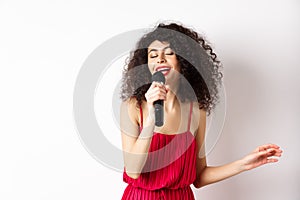 Happy elegant woman in red dress performing with microphone, singing karaoke and smiling, standing over white background