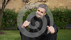 Happy elegant young businessman smiling gesturing thumb up looking at camera sitting outdoors in sunshine. Front view
