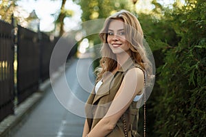 Happy elegant young woman in trendy summer dress with stylish black leather handbag posing on the street near green trees