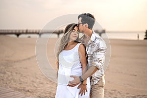 Happy embraced couple at beach. Handsome man hugging his pregnant wife and enjoying together tranquil moments at sunset.