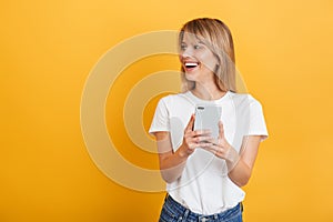 Happy emotional young blonde woman posing isolated over yellow wall background dressed in white casual t-shirt using mobile phone