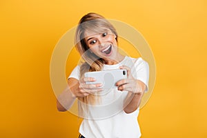 Happy emotional young blonde woman posing isolated over yellow wall background dressed in white casual t-shirt using mobile phone