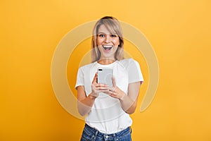 Happy emotional young blonde woman posing isolated over yellow wall background dressed in white casual t-shirt using mobile phone