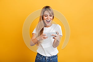 Happy emotional young blonde woman posing isolated over yellow wall background dressed in white casual t-shirt using mobile phone