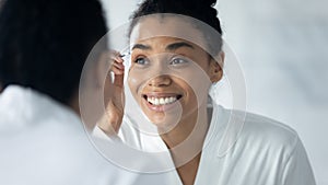 Happy engaged Black millennial girl in bathrobe plucking eyebrows