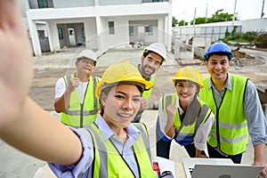 Happy engineers and formats, male and female colleagues in safety vests with helmets taking selfies at under-construction building