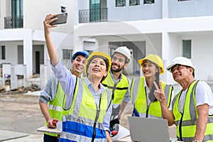 Happy engineers and formats, male and female colleagues in safety vests with helmets taking selfies at under-construction building