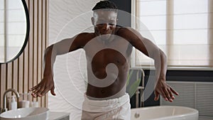 Happy ethnic African American man with bath towel on hips dancing shirtless at bathroom hotel funny naked handsome