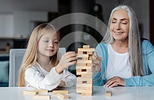 Happy european little girl and old woman play in tower game on table in living room interior