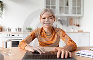 Happy european teenage girl blonde typing on keyboard by laptop in cozy kitchen interior