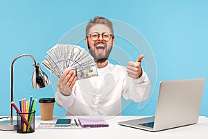 Happy excited bearded man showing thumbs up like gesture holding fan of dollar cash sitting at workplace with laptop, easy profit