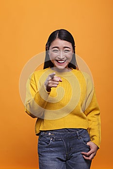 Happy excited beautiful woman wearing yellow sweater pointing index finger at camera on orange background.