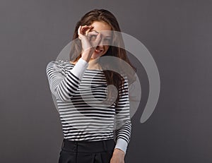 Happy excited business brunette woman doing spyglass sign looking in fingers near the eye in stripped casual t-shirt. Concept