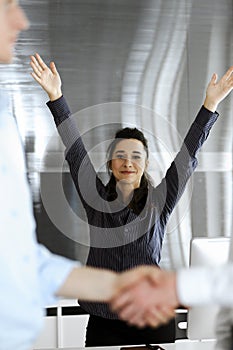 Happy and excited business woman raising hands because of success agreement at meeting. Two men shaking hands in office
