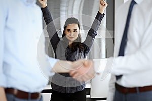 Happy and excited business woman raising hands because of success agreement at meeting. Two men shaking hands in office