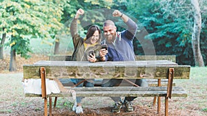 Happy excited couple man and woman sitting in autumn garden with cat, looking to their phones and screaming