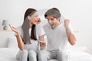 Happy excited emotional young caucasian man and woman hold pregnancy test in bedroom interior and rejoice