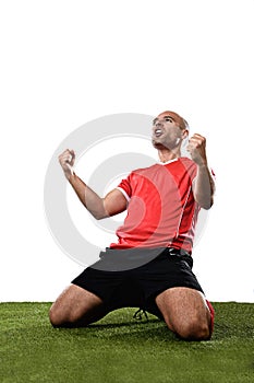 Happy and excited football player in red jersey celebrating scoring goal kneeling on grass pitch