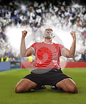 Happy and excited football player in red jersey celebrating scoring goal kneeling on grass pitch