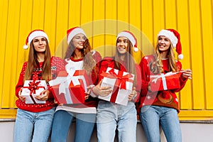 Happy excited group of friends dressed in red sweaters and santa claus hats holding christmas presents while standing near yellow