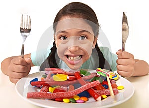 Happy excited Latin female child holding fork and knife sitting at table ready for eat a dish full of candy