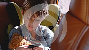 Happy excited little 4-6 year old Caucasian boy smiling, using smartphone sitting in car child safety seat with a toy.