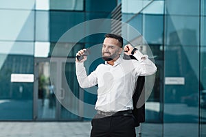 Happy excited mature businessman celebrate victory with fists raised in the air staying against modern glass office