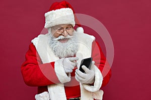 Happy excited Santa Claus using smart phone isolated on red background.