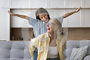 Happy excited strong grandma piggybacking granddaughter, sitting on home sofa photo