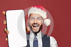 Happy excited successful businessman in red Santa hat showing smartphone with blank empty white screen display for new App