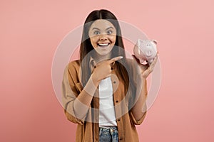 Happy excited teen girl pointing at piggy bank and looking at camera