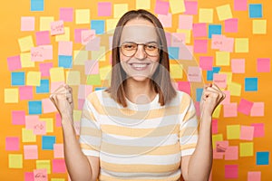 Happy excited woman with brown hair standing isolated over yellow background with stickers, celebrating her winning, clenched