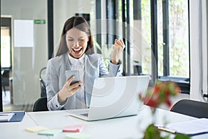 Happy excited woman holding phone and triumphing with fist up in office