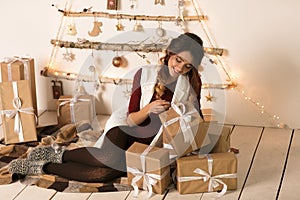 Happy excited woman sitting near the rustic Christmas tree with many Christmas gifts over white backgound