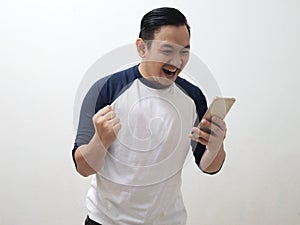 Happy excited young Asian man smiling shouting and  shows winning gesture when looking at his smart phone, receiving good news