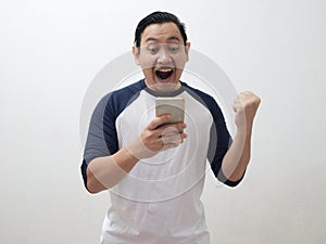 Happy excited young Asian man smiling shouting and  shows winning gesture when looking at his smart phone, receiving good news